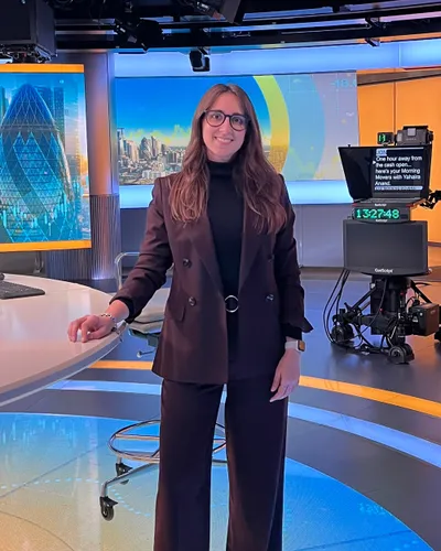 Maria Pastor in a burgundy trouser suit, smiling in a television news studio