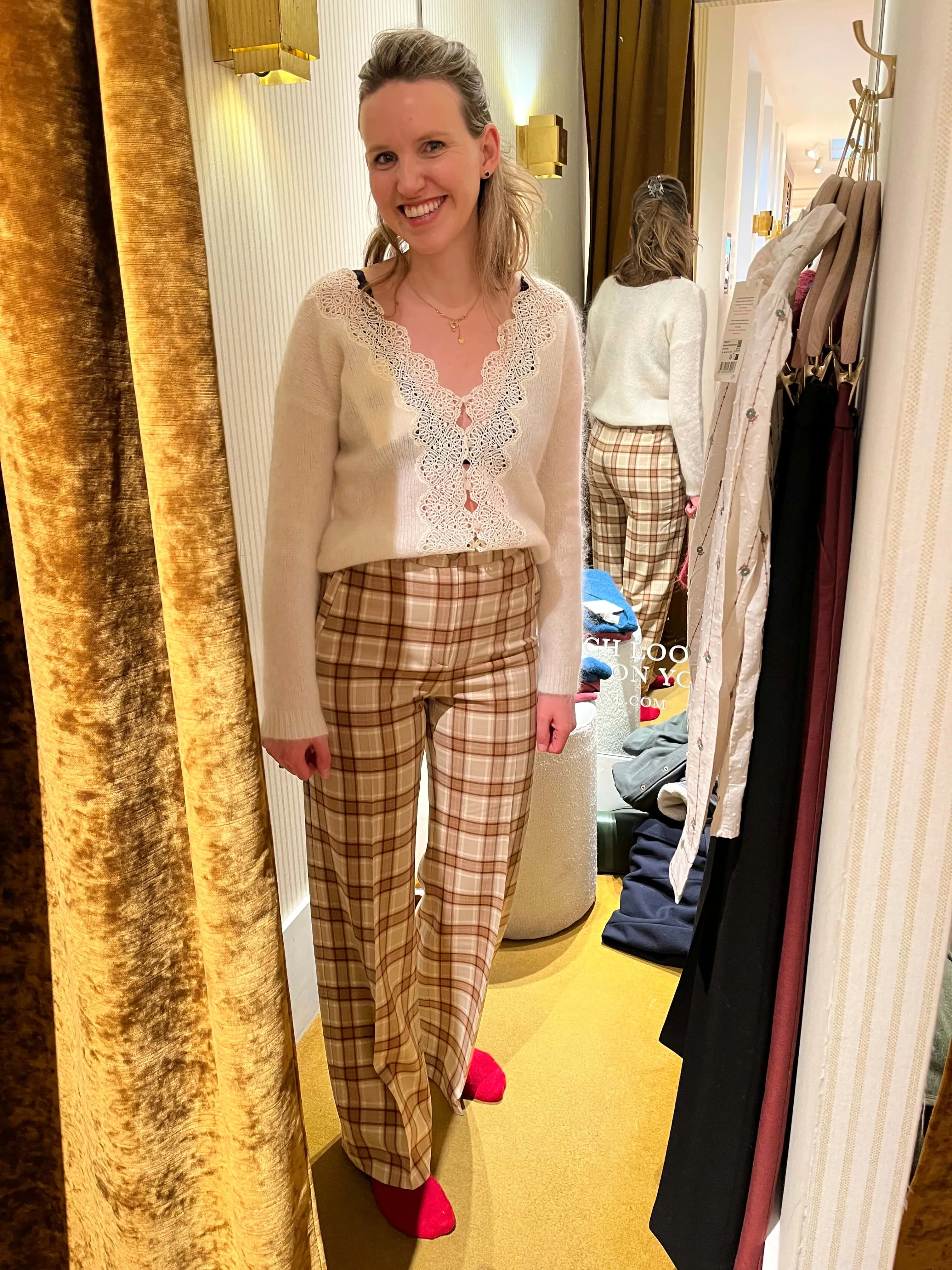 A smiling client in a fitting room wearing a cream lace-trimmed cardigan and check trousers