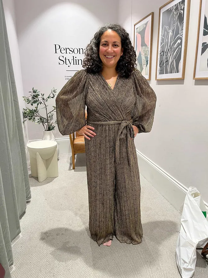 A smiling client in a fitting room wearing a belted patterned jumpsuit