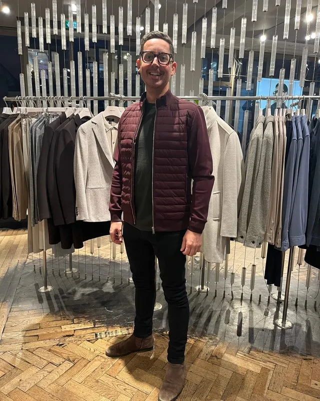 A smiling client in a burgundy quilted gilet over a dark jumper beside a rail of men's jackets