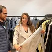 Maria Pastor holding up a pale coat for a male client in front of rails of menswear