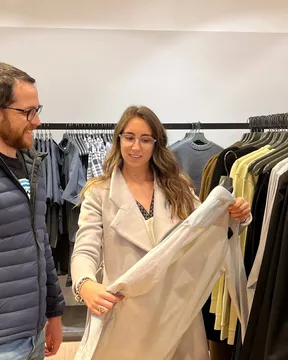 Maria Pastor holding up a pale coat for a male client in front of rails of menswear