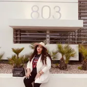 A woman in a white blazer and straw hat sitting against a white wall with a black handbag