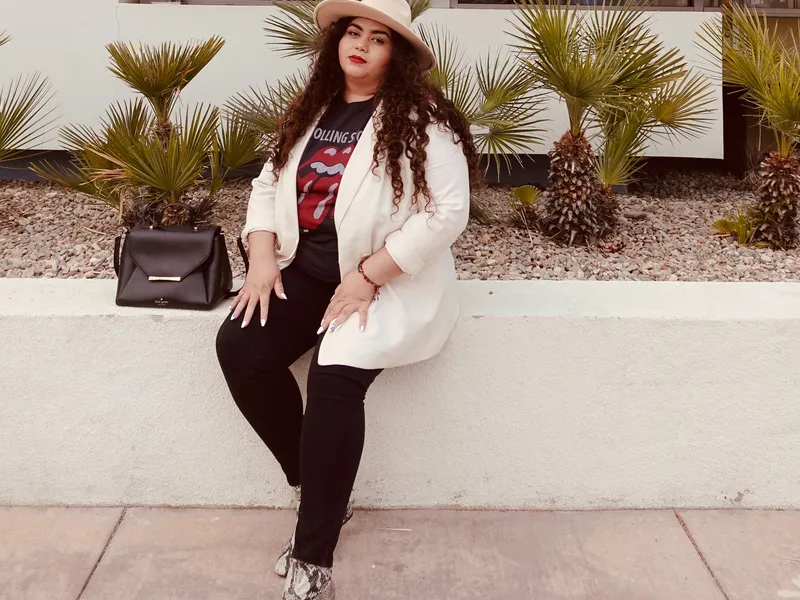 Woman in a white blazer and wide-brimmed hat sitting on a low wall outside a sunlit house