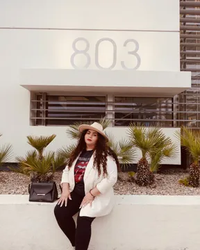 A woman in a white blazer and straw hat sitting against a white wall with a black handbag