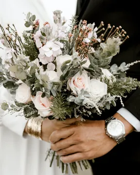 A bride and groom holding a blush and white bouquet between them, her wrist in a gold cuff, his in a dark suit sleeve