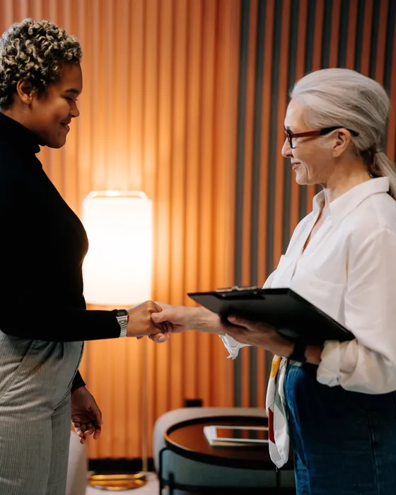 Two women shaking hands in a warm-lit lounge, one holding a clipboard, in front of an orange slatted wall
