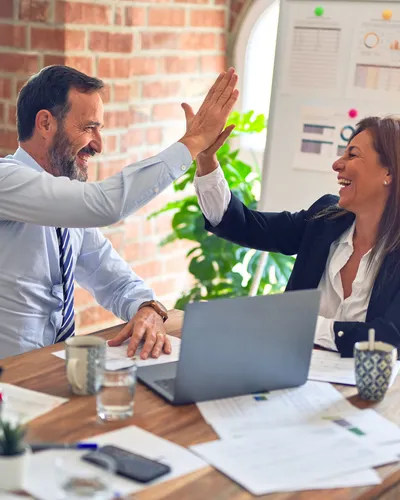 Two colleagues in business dress sharing a high five across a meeting table