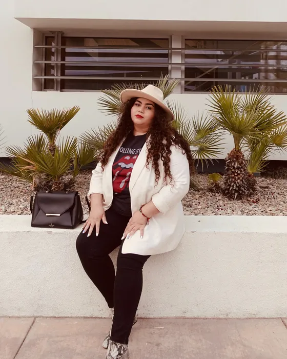 A woman in a white blazer and wide-brimmed hat sitting on a low wall in front of a white building with palms