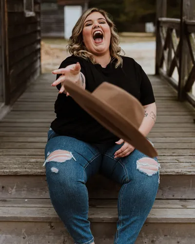 A woman in a black T-shirt and jeans laughing as she tosses a brown hat, sitting on wooden steps