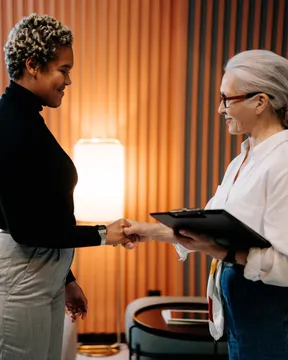 Two women shaking hands in a warm-lit lounge, one holding a clipboard