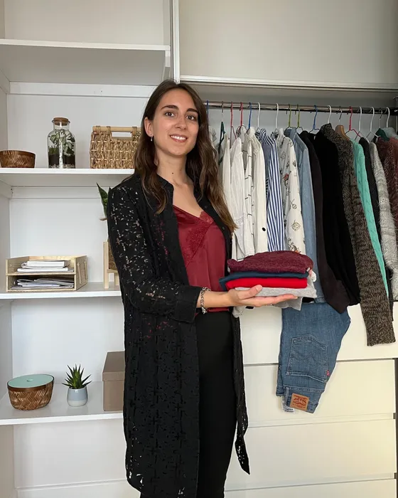 Maria Pastor, Personal Shopper & Stylist, holding a stack of folded clothes by an open wardrobe