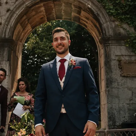 A groom in a navy three-piece suit and burgundy tie, standing beneath a stone archway